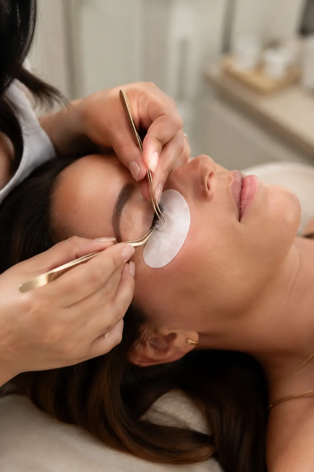 Lash artist applying extensions with gold tweezers and a white gel patch under the eye